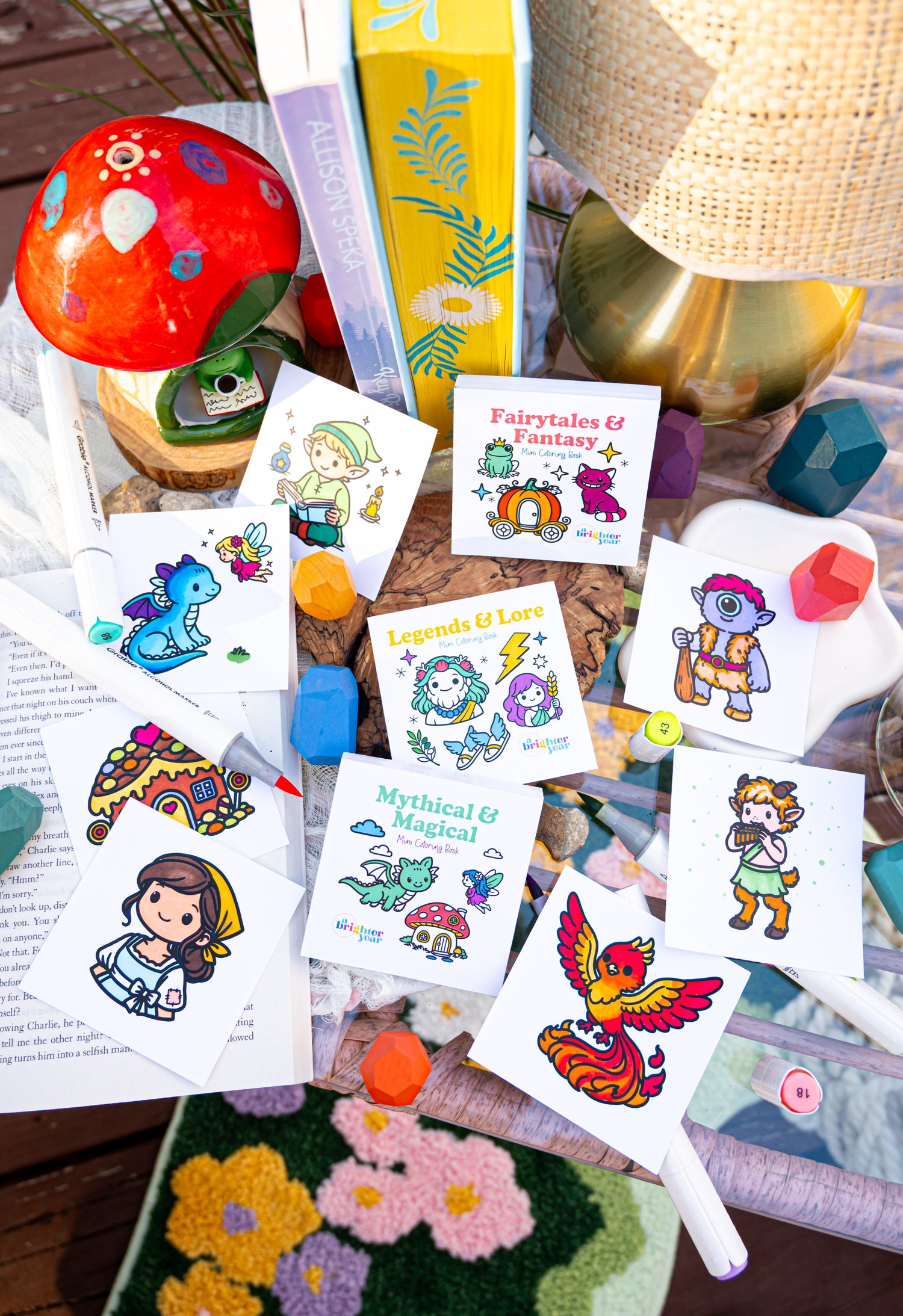 A styled flat-lay of a color your own tarot card deck with multiple illustrated tarot cards spread out on a light wood surface, alongside coloring markers, a small black bird figurine, a white dish, and translucent orange glassware.