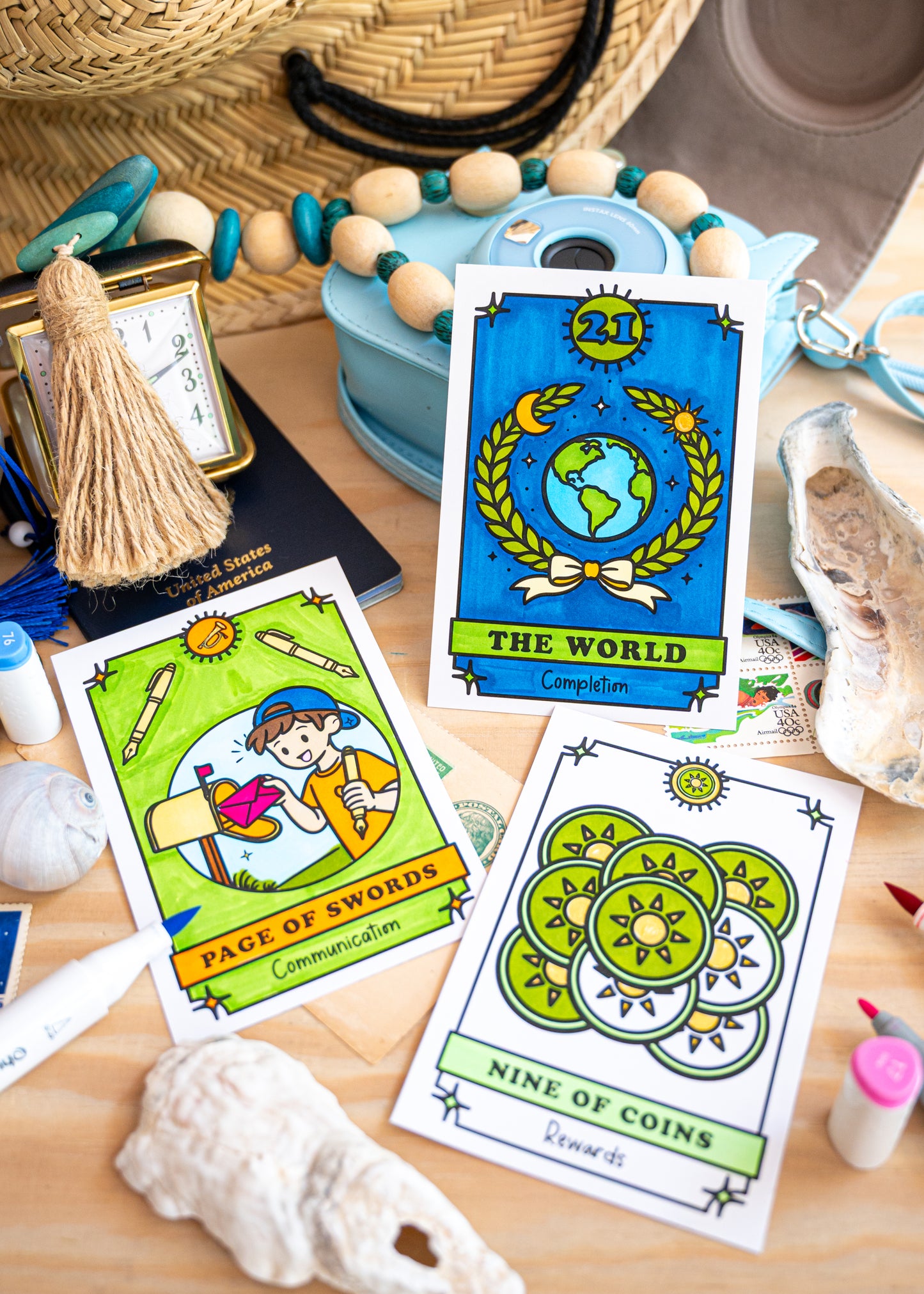 Three illustrated color your own tarot cards are arranged on a wooden surface with beach and craft props around them, including a straw hat, beads, a small clock, a tassel, shells, and paintbrushes. The visible cards read "The World" and "Nine of Coins" with colorful fantasy-style artwork form the brand A Brighter Year. 