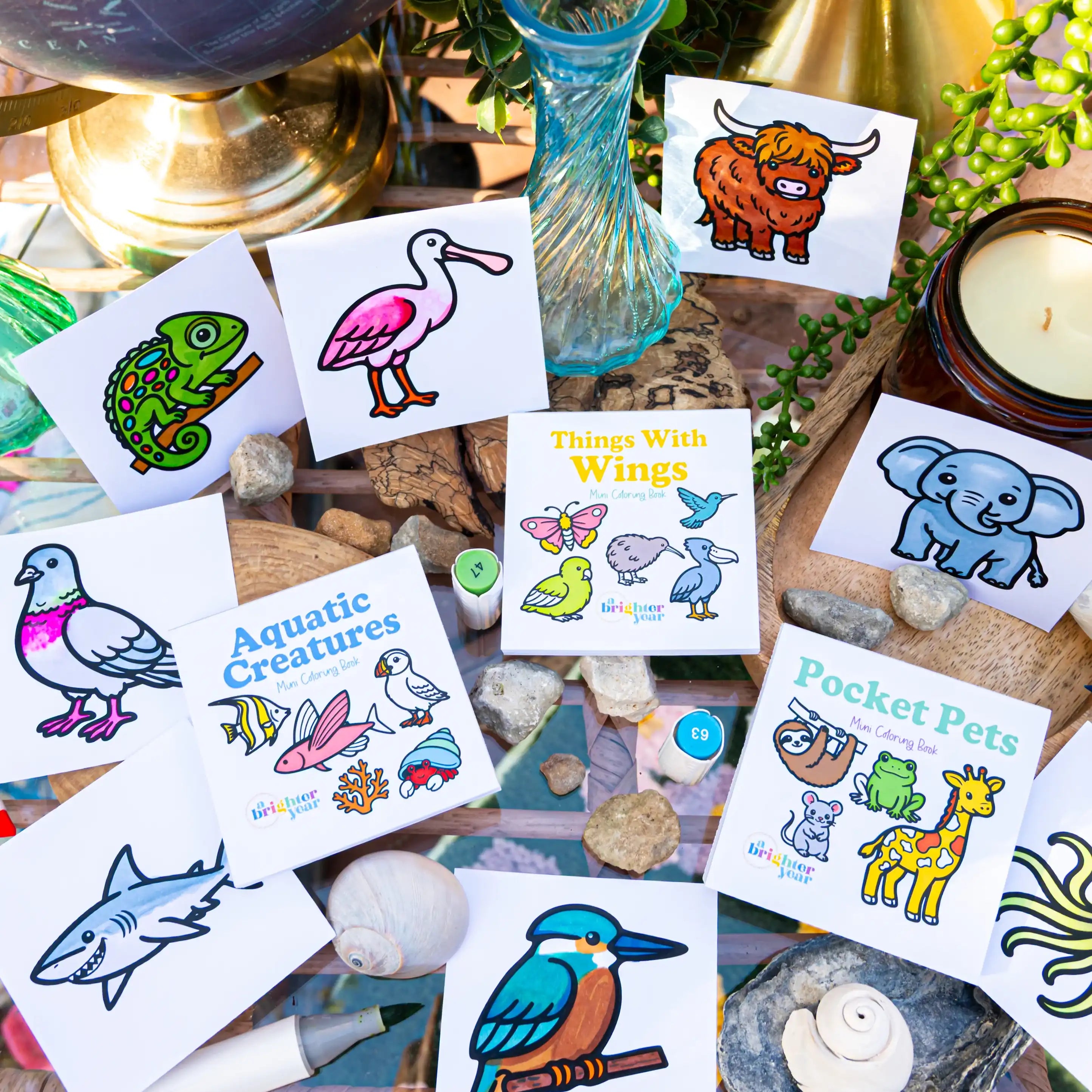 A styled flat-lay of bold and simple coloring books featuring  animals and nature-themed coloring book pages, arranged on rocks with a blue glass vase, candle, and greenery in the background.