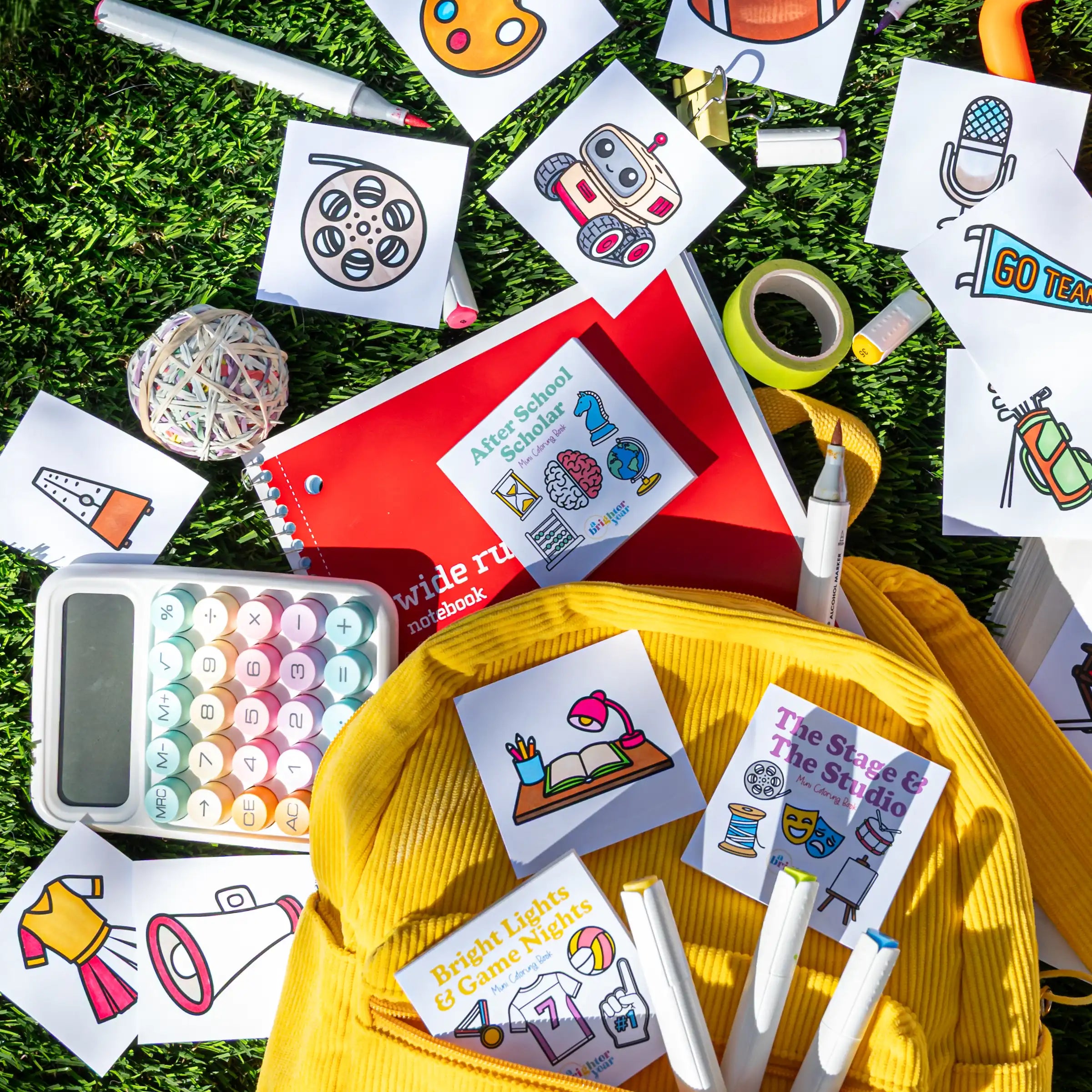 A bright yellow corduroy backpack lies on green grass surrounded by school-themed bold and simple coloring book pages, including markers, a red notebook, tape, and mini coloring books school themed illustrations and titles like 'After School Scholar' and 'The Stage & The Studio', and "Bright Lights and Game Nights".