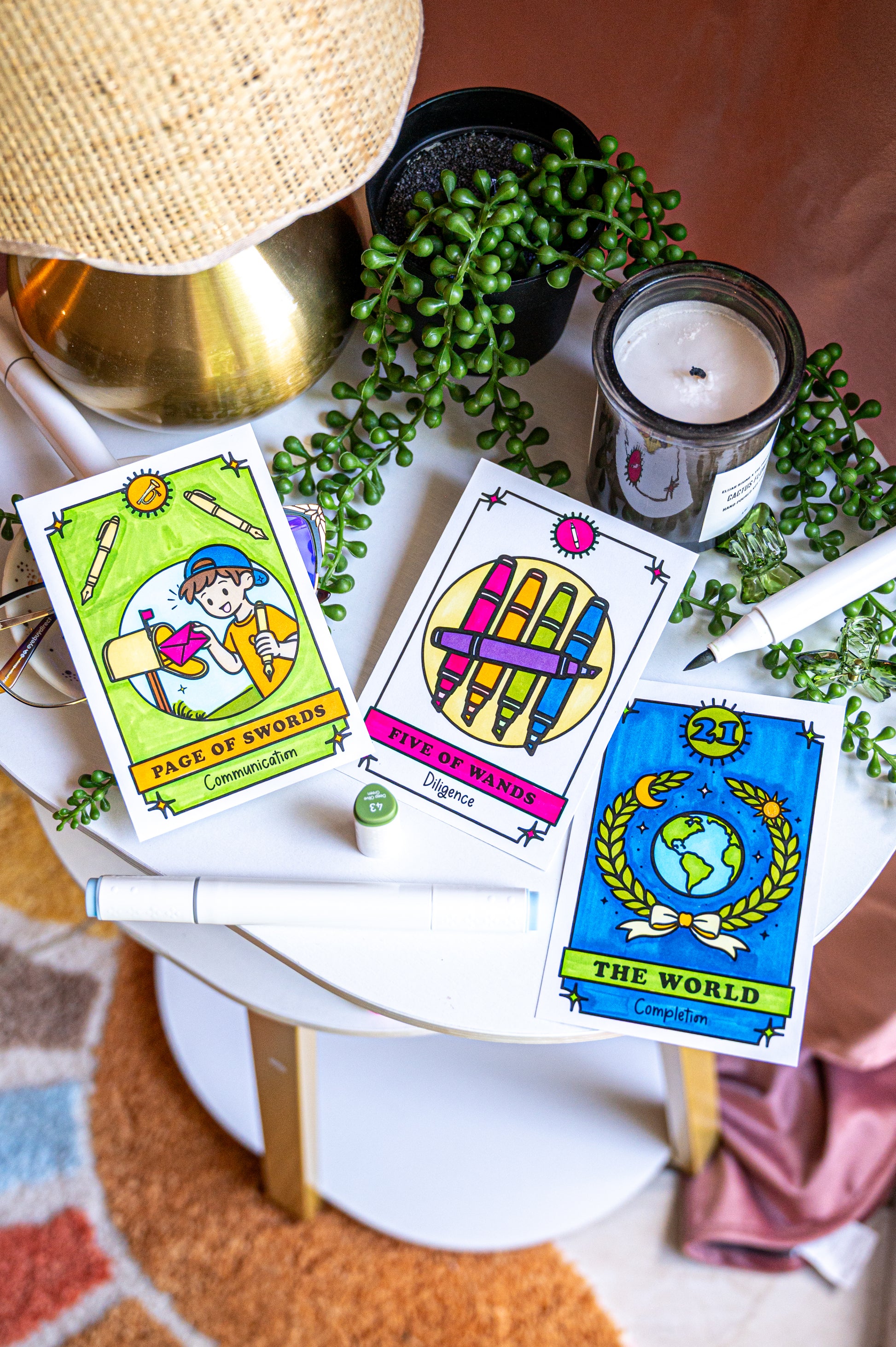 Three color your own tarot-style cards are arranged on a small white side table with a candle, green trailing plant, lamp base, and fabric around them. The cards show colorful fantasy illustrations and titles including 'Page of Swords,' 'Eight of Wands,' and 'The World.