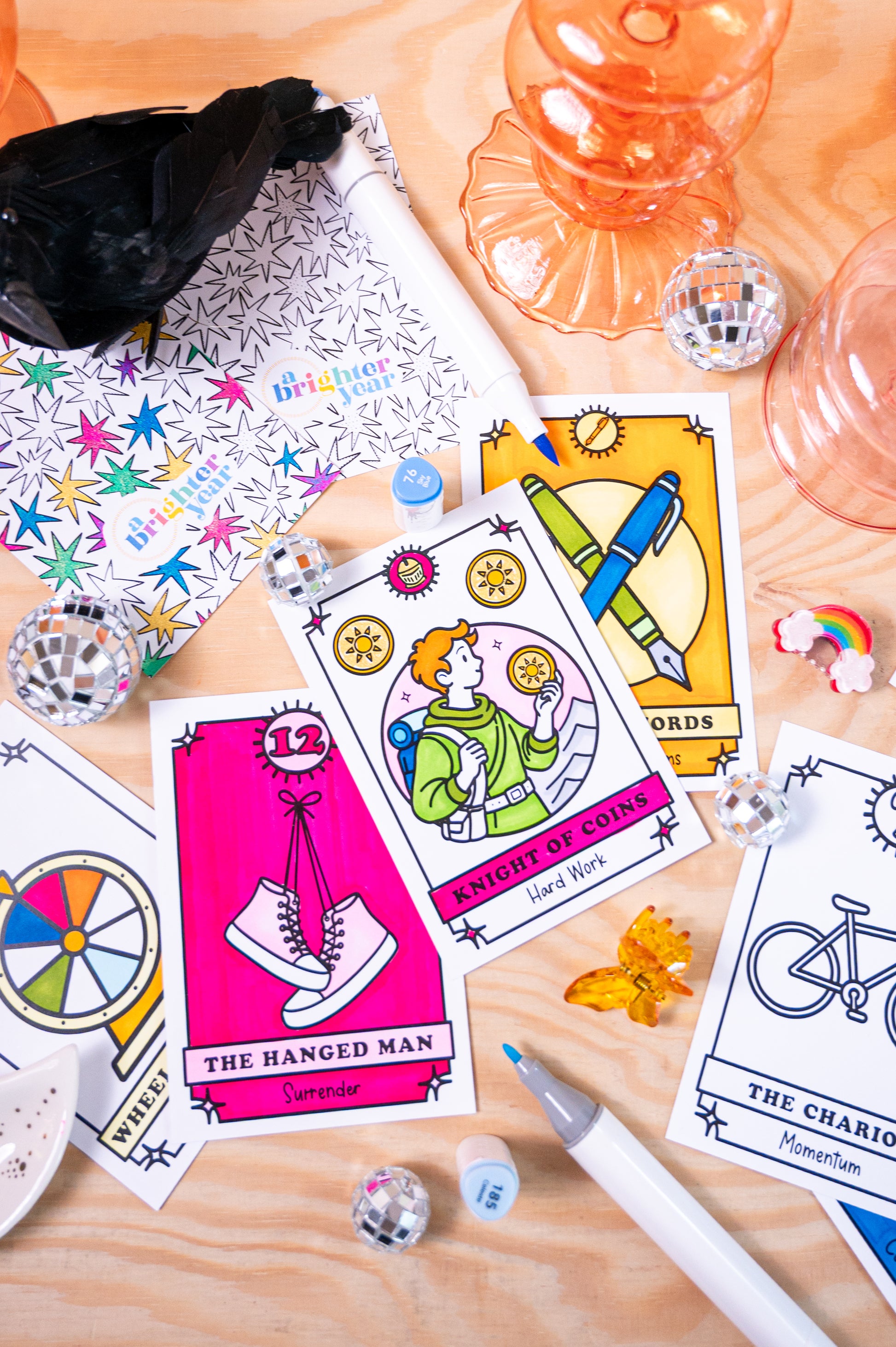 A styled flat-lay of a tarot card deck on a light wood surface, featuring color your own illustrated cards such as 'The Hanged Man', 'Knight of Cups', and 'The Chariot', alongside a patterned box, mini mirrors, a pen, and decorative props from the brand A Brighter Year. 