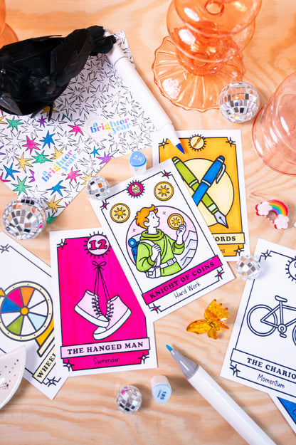 A styled flat-lay of a tarot card deck on a light wood surface, featuring color your own illustrated cards such as 'The Hanged Man', 'Knight of Cups', and 'The Chariot', alongside a patterned box, mini mirrors, a pen, and decorative props from the brand A Brighter Year. 