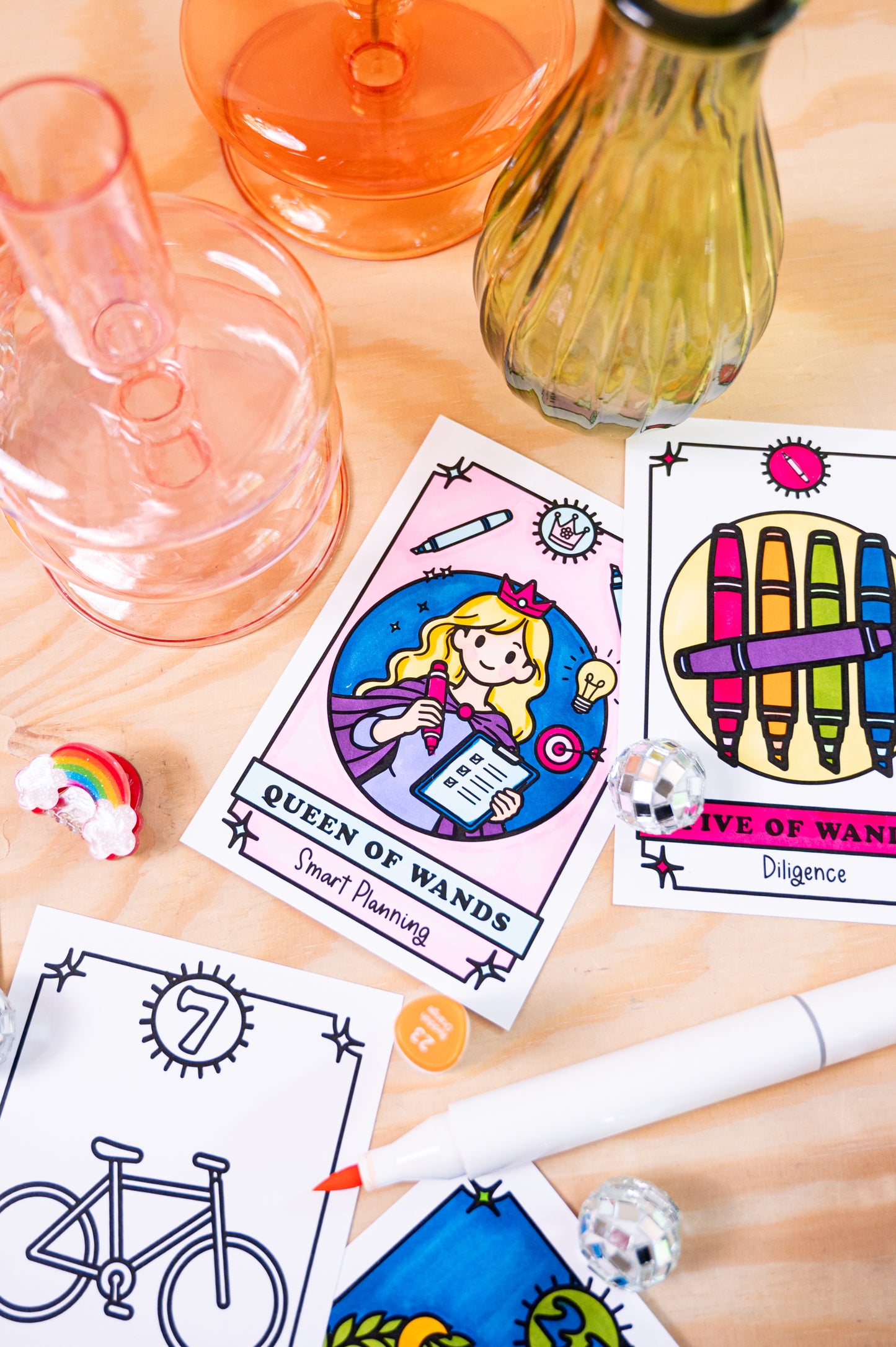 A styled flat-lay of color your own tarot cards on a light wood surface, surrounded by decorative glass containers, a rainbow figurine, and a white marker. The visible cards include Queen of Wands and Five of Wands with colorful illustrated designs.