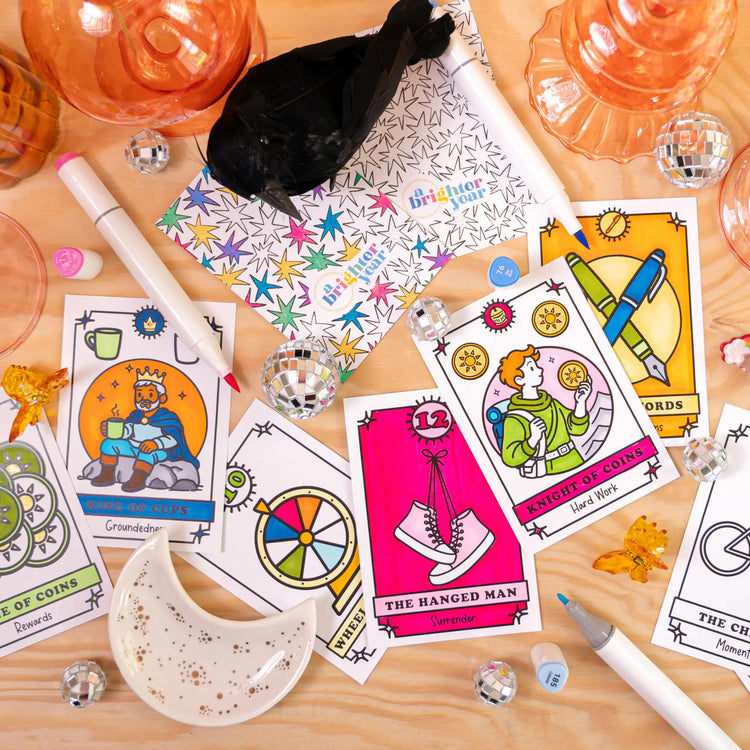 A styled flat-lay of a color your own tarot card deck with multiple illustrated tarot cards spread out on a light wood surface, alongside coloring markers, a small black bird figurine, a white dish, and translucent orange glassware.