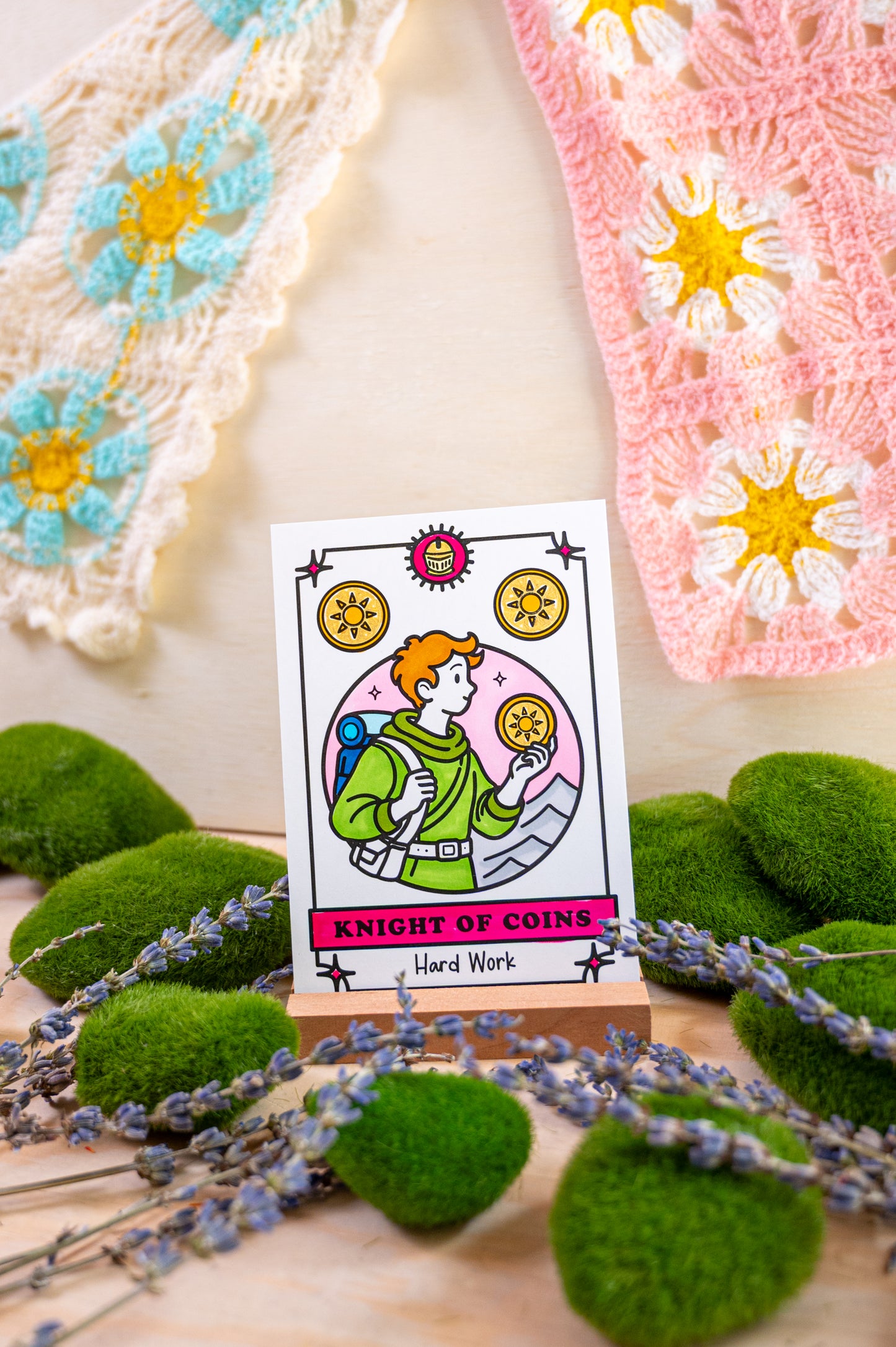 A tarot card reading 'Knight of Coins' and 'Hard Work' displayed on a small wooden stand, surrounded by green moss balls, lavender sprigs, and crocheted textile pieces in the background.