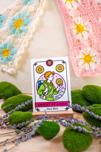 A tarot card reading 'Knight of Coins' and 'Hard Work' displayed on a small wooden stand, surrounded by green moss balls, lavender sprigs, and crocheted textile pieces in the background.
