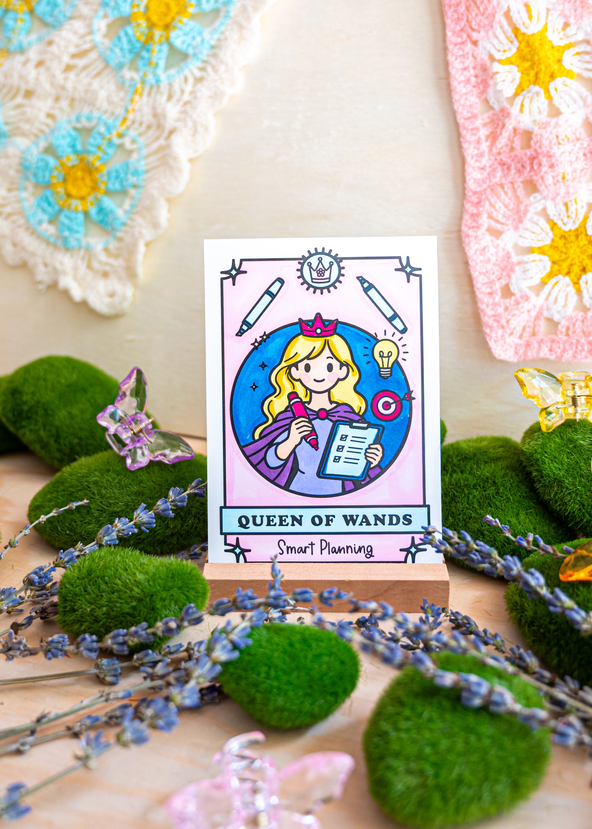 A tarot-style card reading 'QUEEN OF WANDS' with a cartoon girl holding a wand, displayed on a small stand among green moss balls, lavender sprigs, butterflies, and crocheted textile pieces in the background.