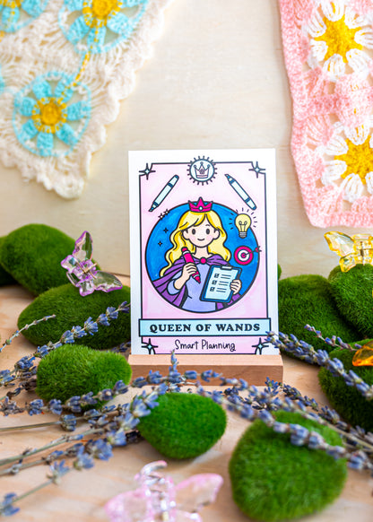 A tarot-style card reading 'QUEEN OF WANDS' with a cartoon girl holding a wand, displayed on a small stand among green moss balls, lavender sprigs, butterflies, and crocheted textile pieces in the background.