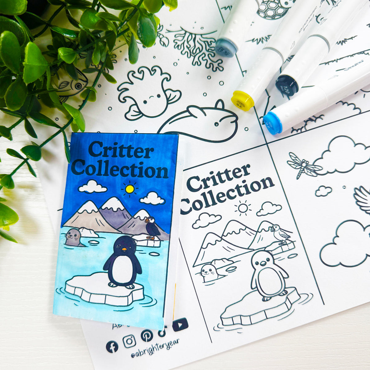 A foldable mini coloring book titled 'Critter Collection' featuring a colorful illustrated cover with a penguin on an iceberg, shown alongside the black-and-white uncolored interior pages and two markers on a white surface with green leaves in the corner.